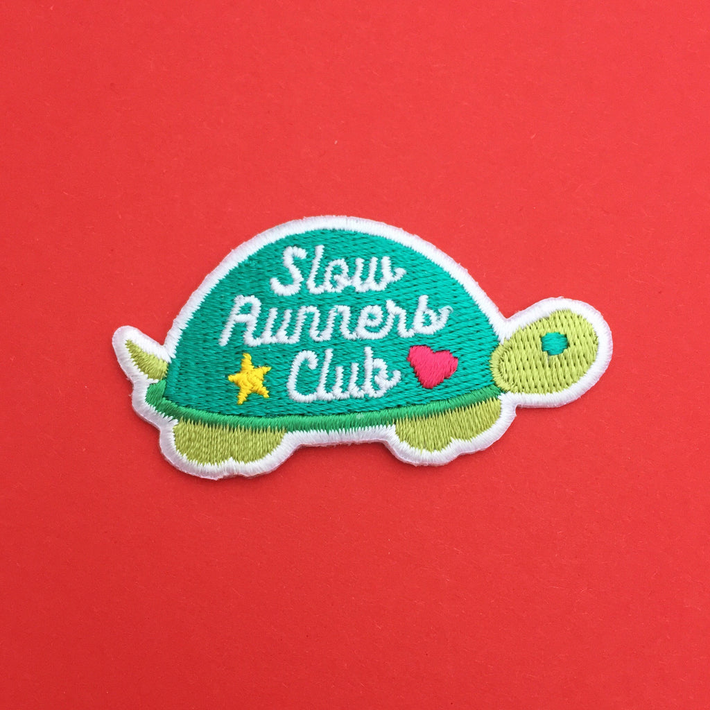 Embroidered patch shaped like a tortoise. The shell is dark green and the head, feet, and tail are light green. On the shell are the words Slow Runners Club in white script decorated with a star and heart. The patch has a white merrowed edge.
