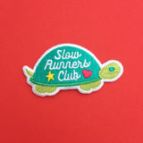 Embroidered patch shaped like a tortoise. The shell is dark green and the head, feet, and tail are light green. On the shell are the words Slow Runners Club in white script decorated with a star and heart. The patch has a white merrowed edge.