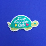 Embroidered patch shaped like a tortoise. The shell is dark green and the head, feet, and tail are light green. On the shell are the words Slow Runners Club in white script decorated with a star and heart. The patch has a white merrowed edge.