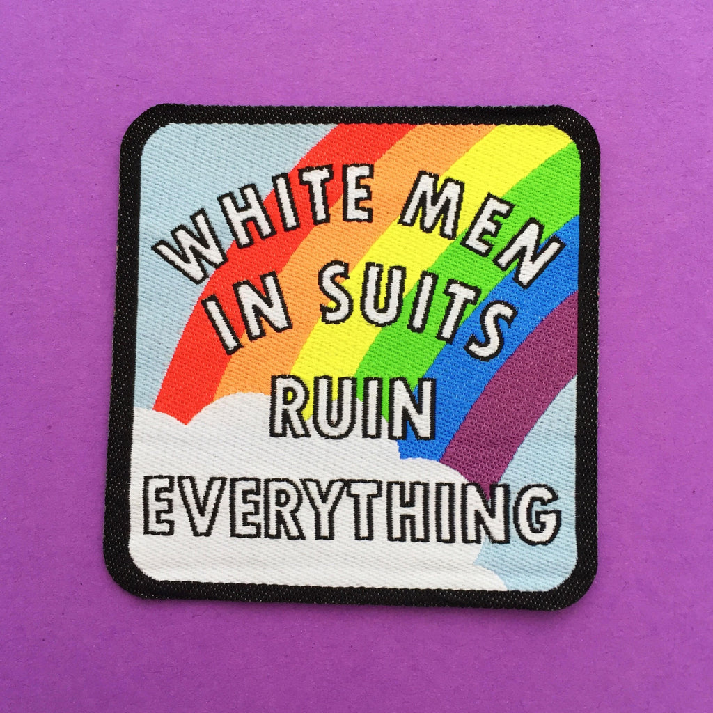 Square woven patch with rounded corners. It reads White Men in Suits Ruin Everything in white capitals. Behind the words a rainbow emerges from a cloud on a pale blue sky. The patch has a black border.