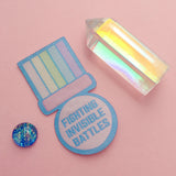 Woven patch shaped like a medal. A pastel-rainbow ribbon section is above a purple bar. Below the bar, the medal is pastel pink, round, and bears the words Fighting Invisible Battles in capital letters. The patch has a blue border.