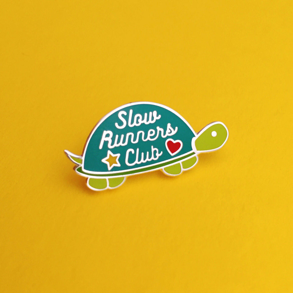 Metal pin shaped like a tortoise. The shell is dark green and the head, feet, and tail are light green. On the shell are the words Slow Runners Club in silver script decorated with a star and heart.