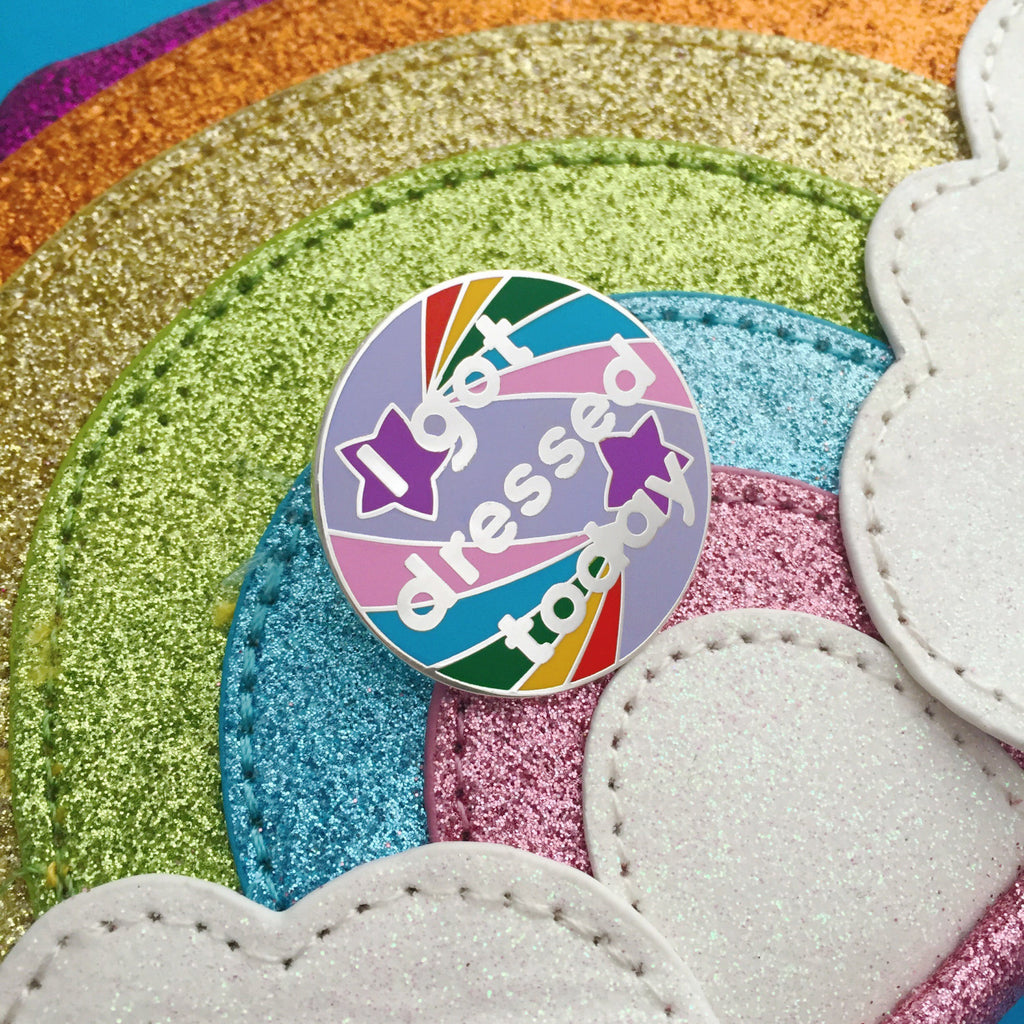 Circular pin that reads I got dressed today in silver letters on lilac enamel. Behind the words are two purple shooting stars with multi-coloured tails. The pin is on a glitter rainbow background.