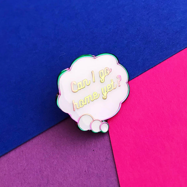 Enamel pin depicting a white, cloud-shaped thought bubble. Inside the bubble is the question Can I Go Home Yet? The metal has a rainbow finish. The pin is shown on a pink, purple, and blue background.