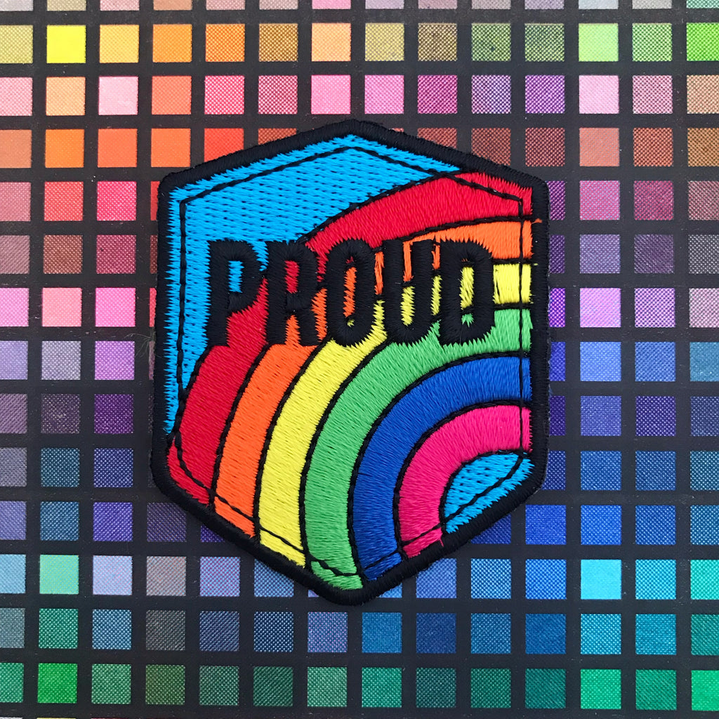 Embroidered patch in a shield shape that reads Proud in black capital letters. The background is a bright rainbow on a blue sky. The patch has black merrowed edges.