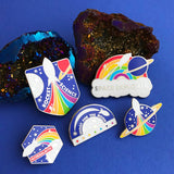 Five metal pins beside dark, sparkly geodes. The pins have various designs each reading individually; Rocket Science, Space Explorer, Intergalactic, and Shoot For The Moon. The last pin simply shows a white rocket with rainbow trails on a ringed planet.