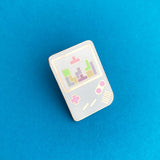 Enamel pin shaped like a light blue Gameboy with pink buttons. The white screen shows a game of tetris in progress with pastel coloured tetrominoes.