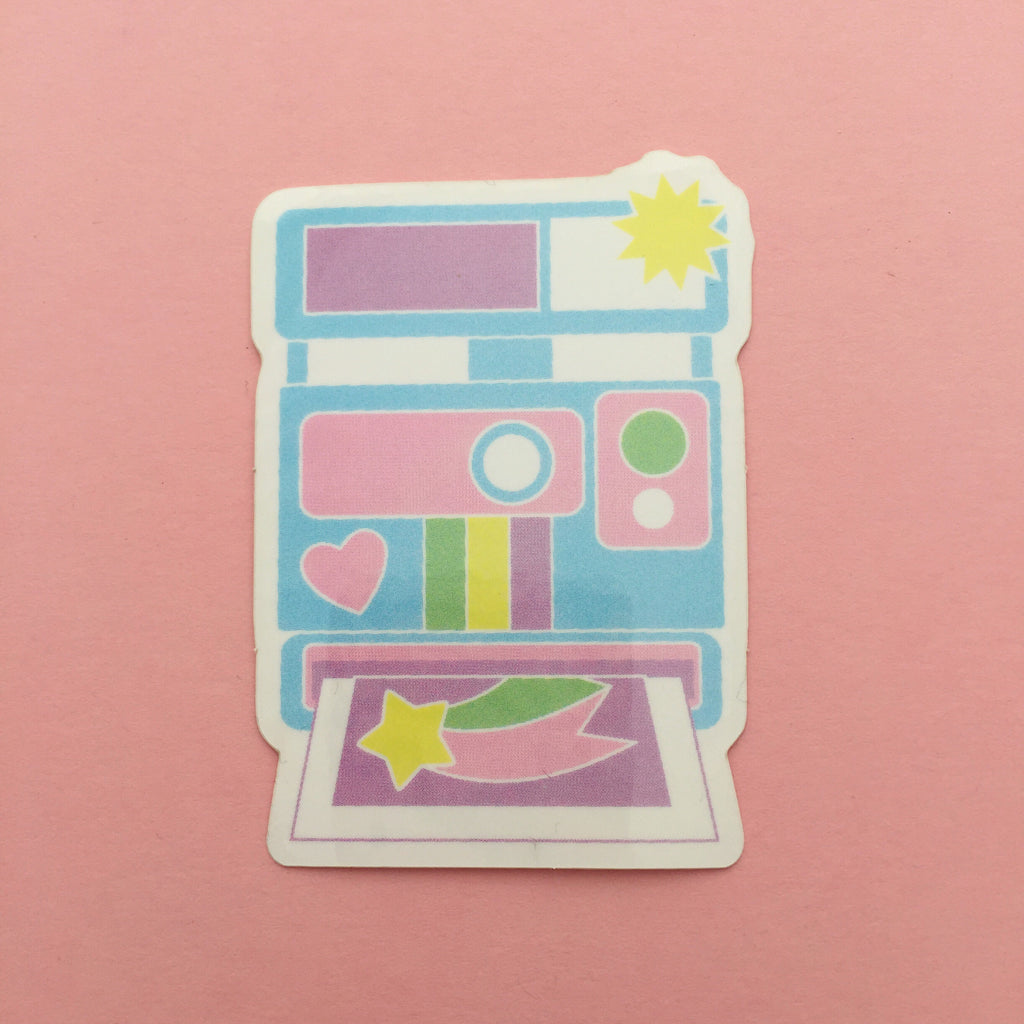 Vinyl sticker shaped like an instant camera in pastel colours. The camera is pastel blue with a heart decoration. The flash has gone off with a yellow starburst and a photo of a shooting star is printing from the bottom. The sticker has a white border.