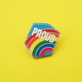 Metal pin in a shield shape that reads Proud in silver capital letters. The background is a bright rainbow on a blue sky.