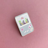Enamel pin shaped like a light blue Gameboy with pink buttons. The white screen shows a game of tetris in progress with pastel coloured tetrominoes.
