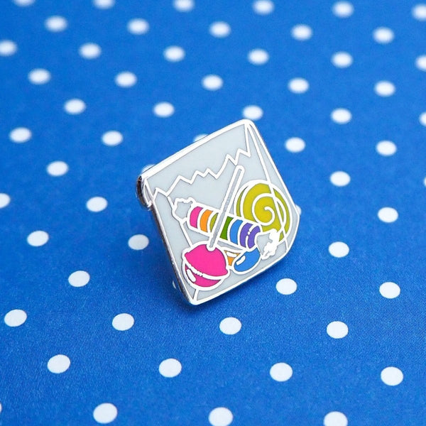 Enamel pin in the shape of a paper bag full of brightly coloured sweets.  Pin is on a blue and white polka dot background.