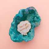 Enamel pin depicting a glittery, cloud-shaped thought bubble. Inside the bubble is the question Can I Go Home Yet? The metal has a rose gold finish. The pin is resting on a turquoise coloured geode crystal.