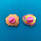 The back of two cloud-shaped enamel pins. One is rose gold, the other is rainbow metal. Both have a pink rubber backing. They are shown on a blue background.