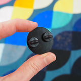 The back of a circular pin being held between a thumb and forefinger. It is black with two black rubber backings.