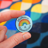 Round, light blue pin with a glitter rainbow in the centre that reads Awkward & Uncoordinated. The pin has a glossy, epoxy layer over the surface. It is being held between a thumb and forefinger.