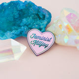 A pink enamel pin in the shape of a heart. The words Feminist Killjoy are written in rainbow-plated metal in a flowing script. Rainbow-plated dots run along the inside border of the heart. The pin is nestled amongst colourful crystals.