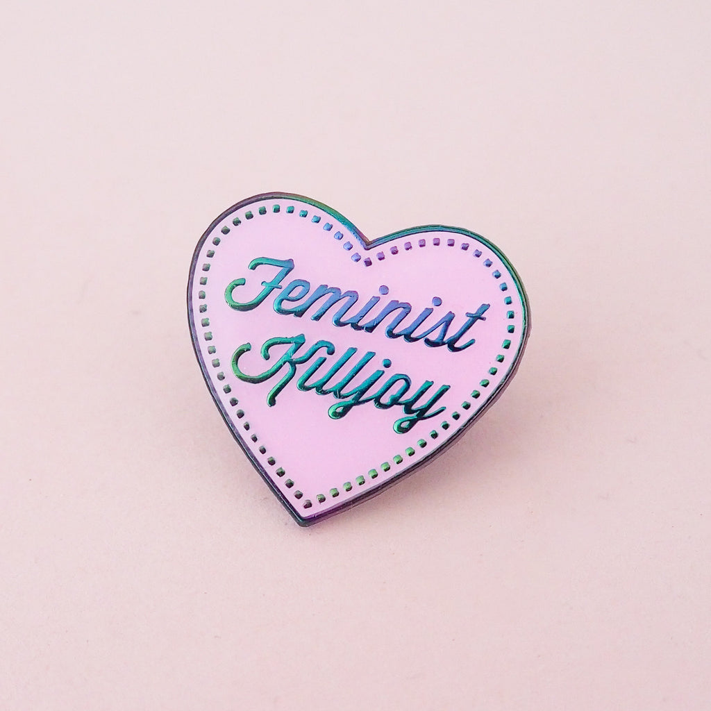 A pink enamel pin in the shape of a heart. The words Feminist Killjoy are written in rainbow-plated metal in a flowing script. Rainbow-plated dots run along the inside border of the heart. The pin is shown on a pink background.