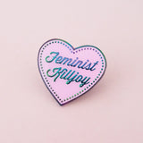 A pink enamel pin in the shape of a heart. The words Feminist Killjoy are written in rainbow-plated metal in a flowing script. Rainbow-plated dots run along the inside border of the heart. The pin is shown on a pink background.