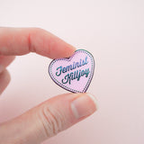 A pink enamel pin in the shape of a heart. The words Feminist Killjoy are written in rainbow-plated metal in a flowing script. Rainbow-plated dots run along the inside border of the heart. The pin is held between a finger and thumb.