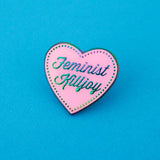 A pink enamel pin in the shape of a heart. The words Feminist Killjoy are written in rainbow-plated metal in a flowing script. Rainbow-plated dots run along the inside border of the heart. The pin is shown on a blue background.