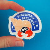 Semi-circular, blue vinyl sticker with the image of a hermit crab in a peach coloured spiral shell. Only the crabs eyes and red claws are visible, peeking out from the darkness of the shell's entrance. The words Exclusively Reclusive are written on the blue section in white capital letters with some white sparkles. The sticker has a white border and is held between a finger and thumb.