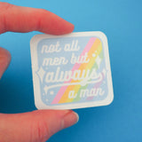 Square vinyl sticker with rounded corners that reads not all men but always a man in white script with decorative white sparkles. The background of the sticker is pale blue with pastel rainbow stripes running diagonally from top-right to bottom-left. The sticker has a white border and is held between a finger and thumb.