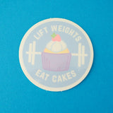 Round vinyl sticker depicting a pastel-coloured cupcake and barbell on a pale blue background. The words Lift Weights Eat Cakes are written in white capitals. The sticker has a white border.