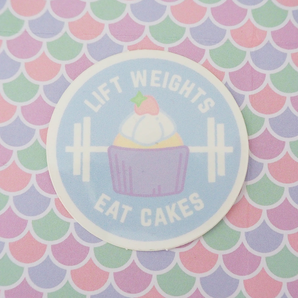 Round vinyl sticker depicting a pastel-coloured cupcake and barbell on a pale blue background. The words Lift Weights Eat Cakes are written in white capitals. The sticker has a white border.