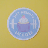 Round vinyl sticker depicting a pastel-coloured cupcake and barbell on a pale blue background. The words Lift Weights Eat Cakes are written in white capitals. The sticker has a white border.