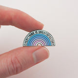 A small enamel pin shaped like a semi-circular rainbow with the text 'EVERYTHING IS UNSATISFACTORY' in black lettering, held between two fingers against a plain light background.