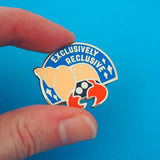Semi-circular, blue enamel pin with the image of a hermit crab in a peach coloured spiral shell. Only the crabs eyes and red claws are visible, peeking out from the darkness of the shell's entrance. The words Exclusively Reclusive are written on the blue section in silver capital letters with some silver sparkles. The pin is being held between a thumb and forefinger in front of a blue background.