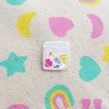 Enamel pin in the shape of a paper bag full of brightly coloured sweets.  Pin is on patterned material featuring hearts, moons, and stars.
