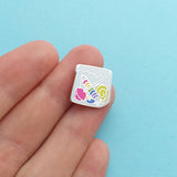 Enamel pin in the shape of a paper bag full of brightly coloured sweets. The pin is shown on a hand for scale, it is roughly the size of a fingertip.