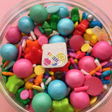 Enamel pin in the shape of a paper bag full of brightly coloured sweets. Pin is in a dish containing sprinkles and shaped candy.
