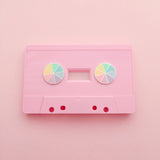 Two round enamel pins, split into eight sections. Each section is filled with a bright rainbow colour, replicating a colour wheel. The pins are sitting in the reels of a pink cassette tape. The tape is on a light pink background.