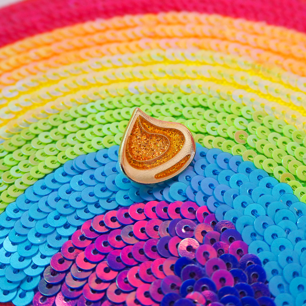 A golden, glittery enamel pin with gold-coloured plating in the shape of a korok seed. The pin is shown on a rainbow-sequinned background.