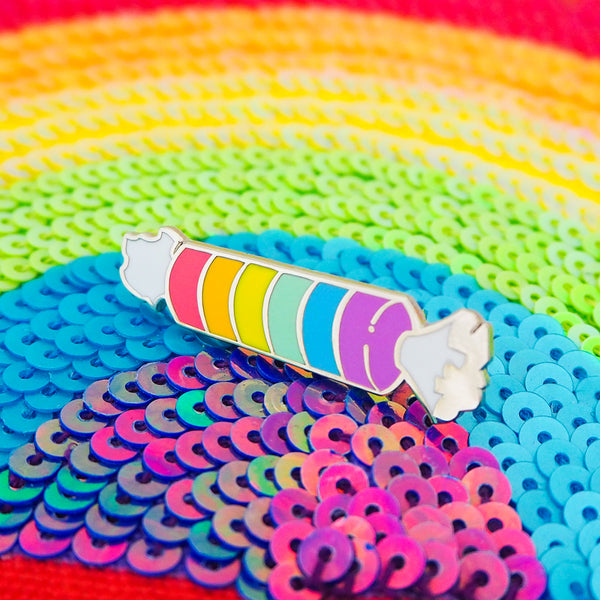 Enamel pin in the shape of a cylindrical pack of fizzy sweets. The sweets are rainbow coloured and the twisted packet ends are white. The pin is lying on a sequinned rainbow background.