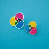Two enamel pins with three overlapping circles. The circles are filled cyan, magenta, and yellow; with the colours mixing in the areas where they overlap. The pin on the left is lightly larger than the other. The pins are on a light blue background.