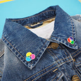 Two pins each on one side of a denim shirt collar. The pins have three overlapping circles. On one pin the circles are filled cyan, magenta, and yellow; and on the other they are red, green and blue. The colours mix in the areas where they overlap.