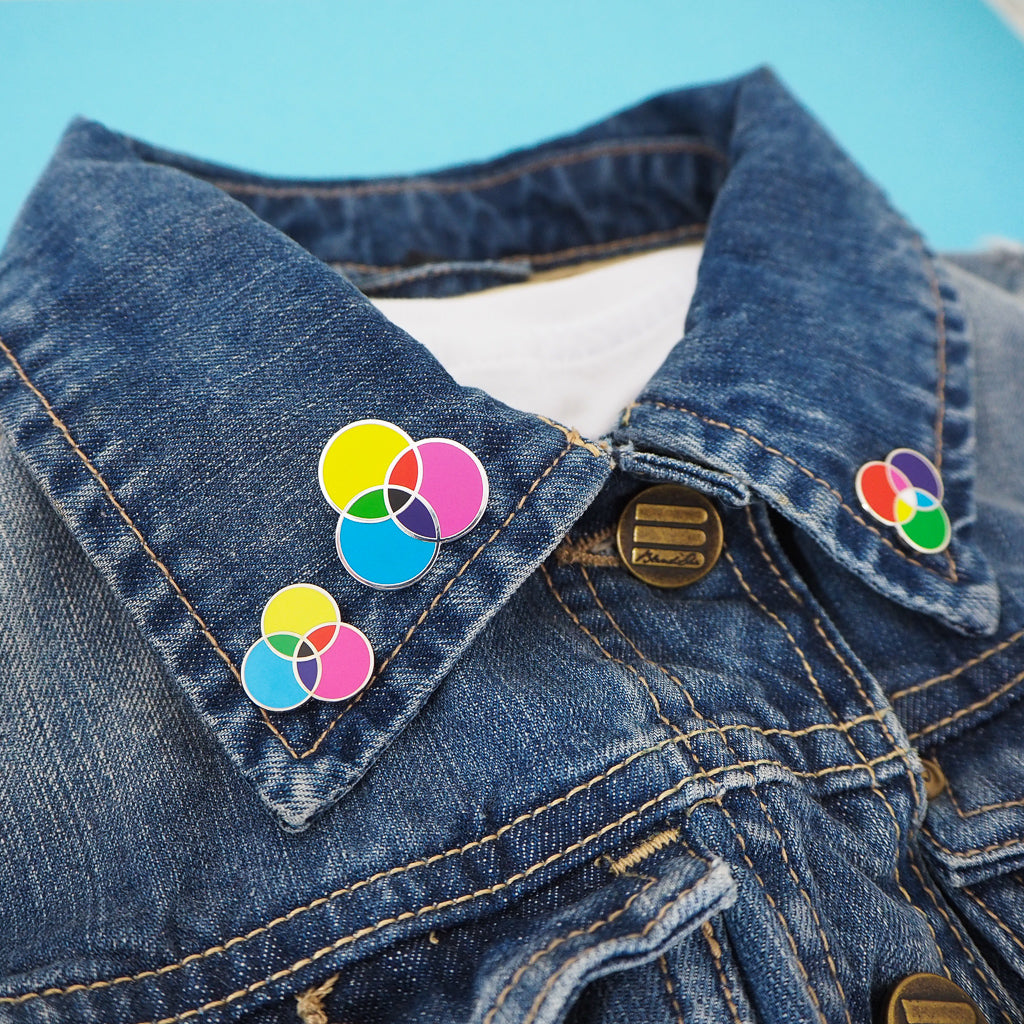 Denim jacket with three pins attached to the collar. The left collar has two enamel pins with three overlapping circles. The circles are filled cyan, magenta, and yellow; with the colours mixing in the areas where they overlap. The pin above and to the right is lightly larger than the other. The pin on the right hand collar is the same design but the colours are red, green, and blue.