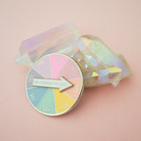 Circular enamel pin divided into eight pastel coloured sections. Each section has the word Yes in capital letters. There is a white arrow that can spin to point to any section with the words Am I Overthinking This? The pin is leaning on a quartz crystal in a light pink background.