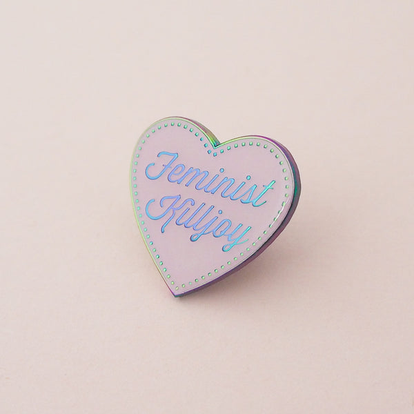 Pastel pink enamel pin in the shape of a heart. The words Feminist Killjoy are written in a flowing script and are rainbow-plated. Rainbow-plated dots run along the inside border of the heart. An epoxy resin coating gives the pin a shiny finish. The pin is shown on a pale pink background.