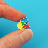 Enamel pin featuring an overlapping circle, square, and triangle. The shapes are brightly coloured, with the colours mixing where they overlap.  The pin is being held between a forefinger and thumb.