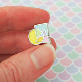 Enamel pin featuring an overlapping circle, square, and triangle. The shapes are pastel coloured, with the colours mixing where they overlap. The pin is being held between a forefinger and thumb.