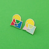 Two enamel pins side by side on a green background. The first pin features an overlapping circle, square, and triangle. The shapes are brightly coloured, with the colours mixing where they overlap. The second pin is the same design in pastel colours.