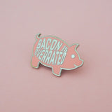 Pink enamel pin in the shape of a pig. On its torso are the word Bacon Is Overrated in capital letters. Pin is on a light pink background.