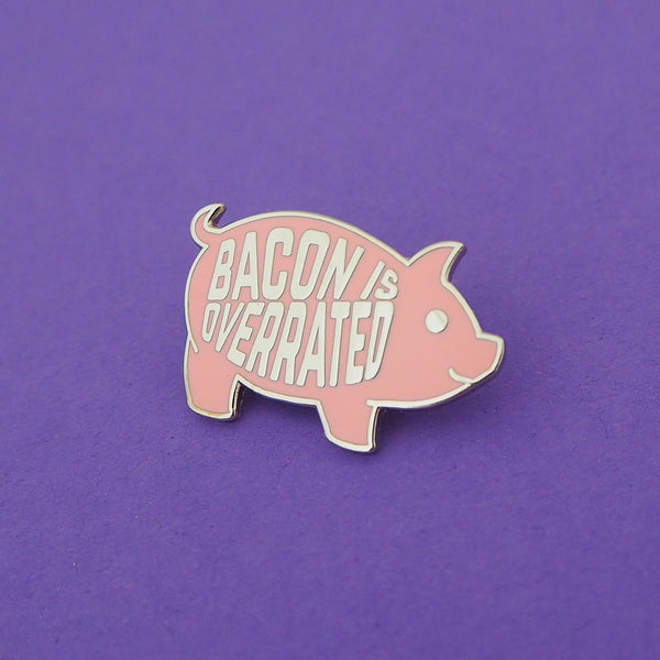 Pink enamel pin in the shape of a pig. On its torso are the word Bacon Is Overrated in capital letters. Pin is shown on a purple background.