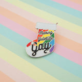 Enamel pin in the shape of a Christmas stocking with rainbow stripes, it reads Making Yuletide Gay in black script. The word Gay is larger and has star sparkles. The pin is on a pastel striped background.