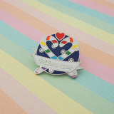 Round enamel pin showing two rainbow striped candy canes crossed at an angle which makes their hooks form a heart shape. There is a small red heart above them and the background is snowflakes on dark blue. There is a ribbon banner at the bottom which reads Christmas Queer. The pin is on a pastel striped background.