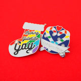Two enamel pins. The first is in the shape of a Christmas stocking with rainbow stripes, it reads Making Yuletide Gay. The word Gay is larger and has star sparkles. The second pin is round, showing two rainbow striped candy canes crossed at an angle which makes their hooks form a heart shape. There is a small red heart above them and the background is snowflakes on dark blue. There is a ribbon banner at the bottom which reads Christmas Queer. The pins are on a bright red background.